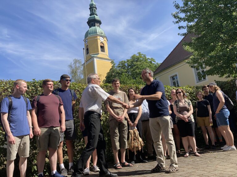 Superintendent Jan Mahling (Jan Malink) begrüßt Fabian Kaulfürst und die Teilnehmer des Sommerkurses für sorbische Sprache und Kultur (im Hintergrund Kirchturm der evangelischen Kirche Königswartha (obersorbisch Rakečanska cyrkej) © Sorbisches Institut (2022)