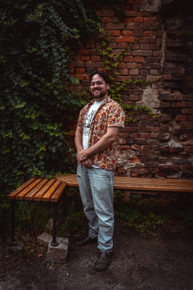 Portrait von Marco, der neben einer Bank vor einem begrünten Mauerwerk steht