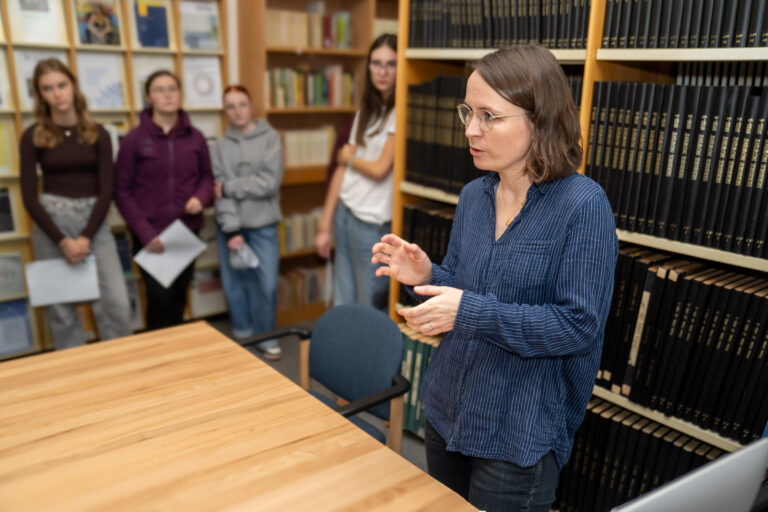 Fachowědomostna referentka Serbskeje centralneje biblioteki steji před knižnej polcu w čitarni Serbskeje centralneje biblioteki, w pozadku steja wjacori šulerjo před wupołoženjom časopisow
