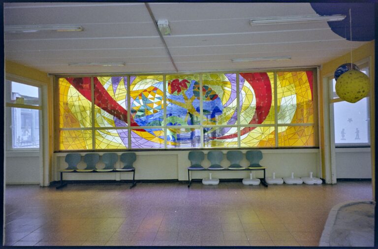 Foto eines bunten wandfüllenden Bleiglasfensters mit Titel ''Mensch im Weltall'' von Jan Buk. Nur bis Brüstungshöhe ist feste Wand mit Stühlen davor. Die großformatige Bleiverglasung wurde im Jahre 1983 von Jan Buck für die damalige Gagarin-Oberschule in Bautzen geschaffen. Abmessungen: 200 cm mal 700 cm.