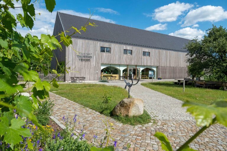 Napohlad Domu tysac hatow w Stróži, prědku we wobrazu dwór z plestrowanym pućom a trawnikom, dale bróžnja, w kotrejž je Dom tysac hatow zaměstnjeny, wyše bróžnje módre njebjo z mróčałkami