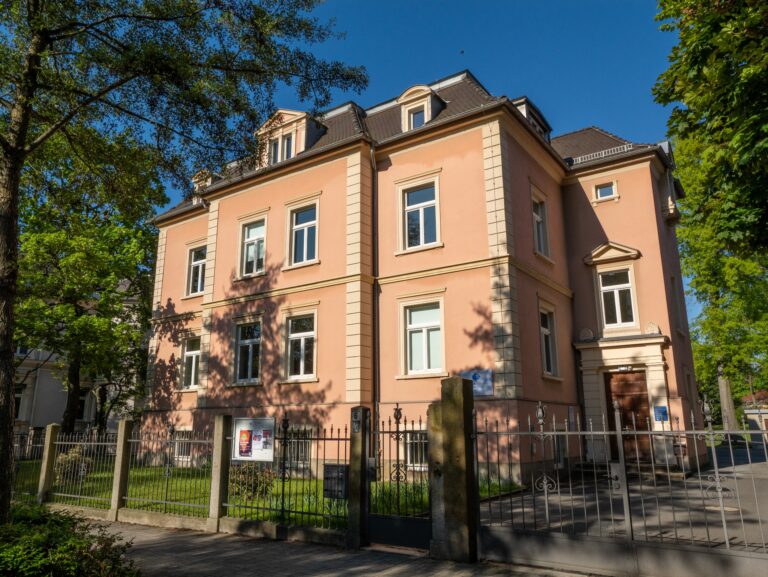 Das Foto zeigt das Gebäude des sorbischen Instituts in Bautzen, an einem sonnigen Tag. Das Bild ist von Gehweg vor dem Institut aufgenommen und zeigt die Vorderseite des Gebäudes mitsamt dem Rasen vor dem Haus, einigen Bäumen und dem Zaun, sowie ein benachbartes Gebäude. Das Haus, eine ehemalige Stadtvilla in der das Sorbische Instut mit einem Anbau im hinteren Teil, der die Bibliothek und das Archiv beinhaltet ist in einem lachsfarbenen Ton gestrichen und erstreckt sich über drei Stockwerke.